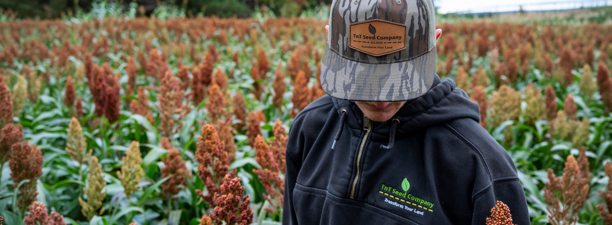 A person wearing a camouflage trucker hat and a black hoodie with green and white logo embroidery stands in a field of red and green plants. The hoodie has a visible 'The Seed Company' logo on the left chest.