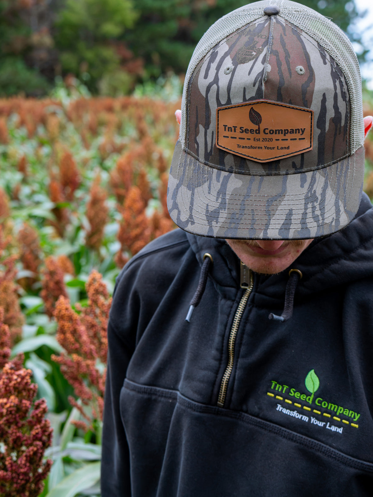 A man wearing a camouflage trucker hat with a 'TnT Seed Company' patch and a matching black pullover hoodie with the 'TnT Seed Company' logo, standing in a field of red sorghum.